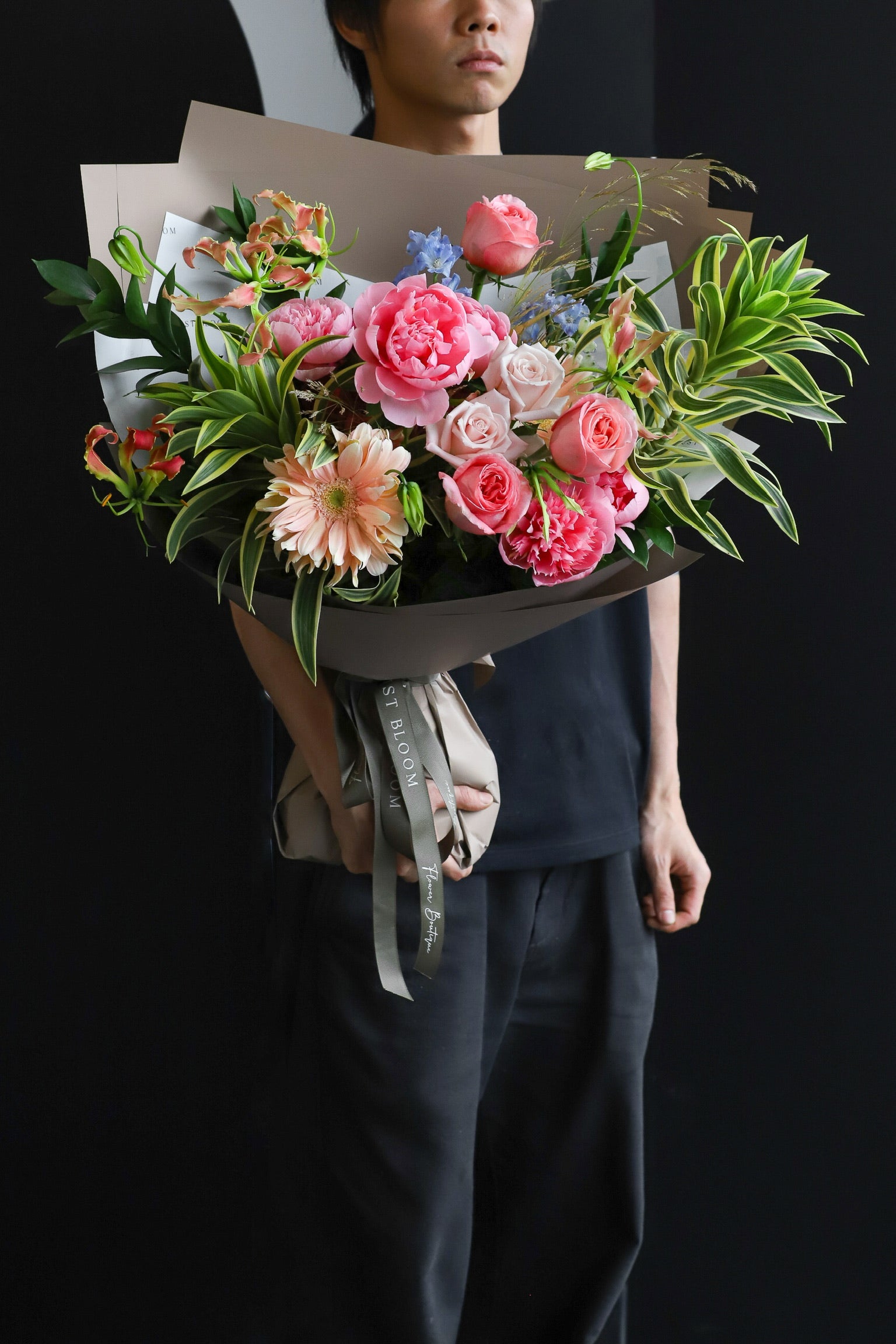  A model in dark clothing holding The Botanist luxury bouquet by Just Bloom Hong Kong, showcasing peonies, roses, gerberas, flame lilies and tropical foliage in taupe wrapping with olive ribbon
