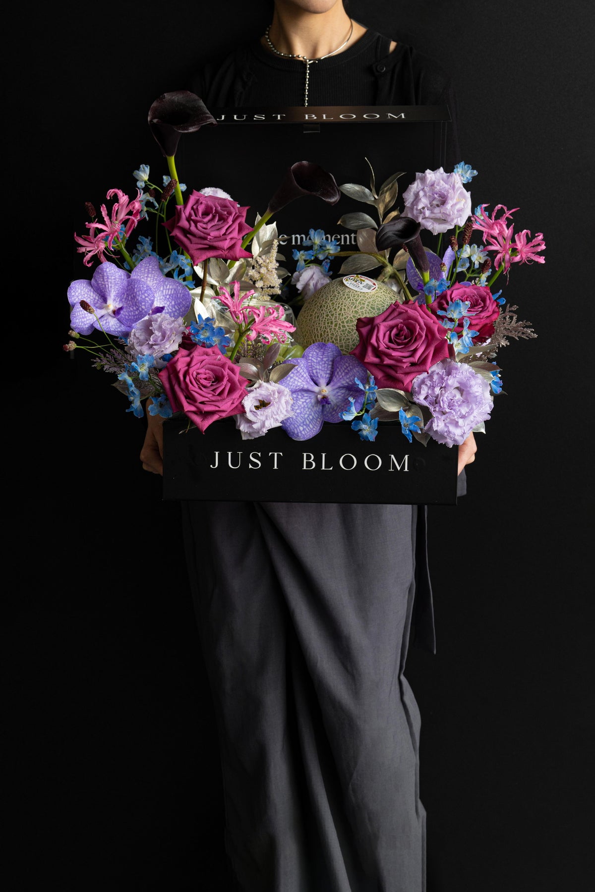 Model holding a large luxury floral hamper in deep purple and magenta tones with premium fruits in a black Just Bloom box against a dark background — Just Bloom Hong Kong
