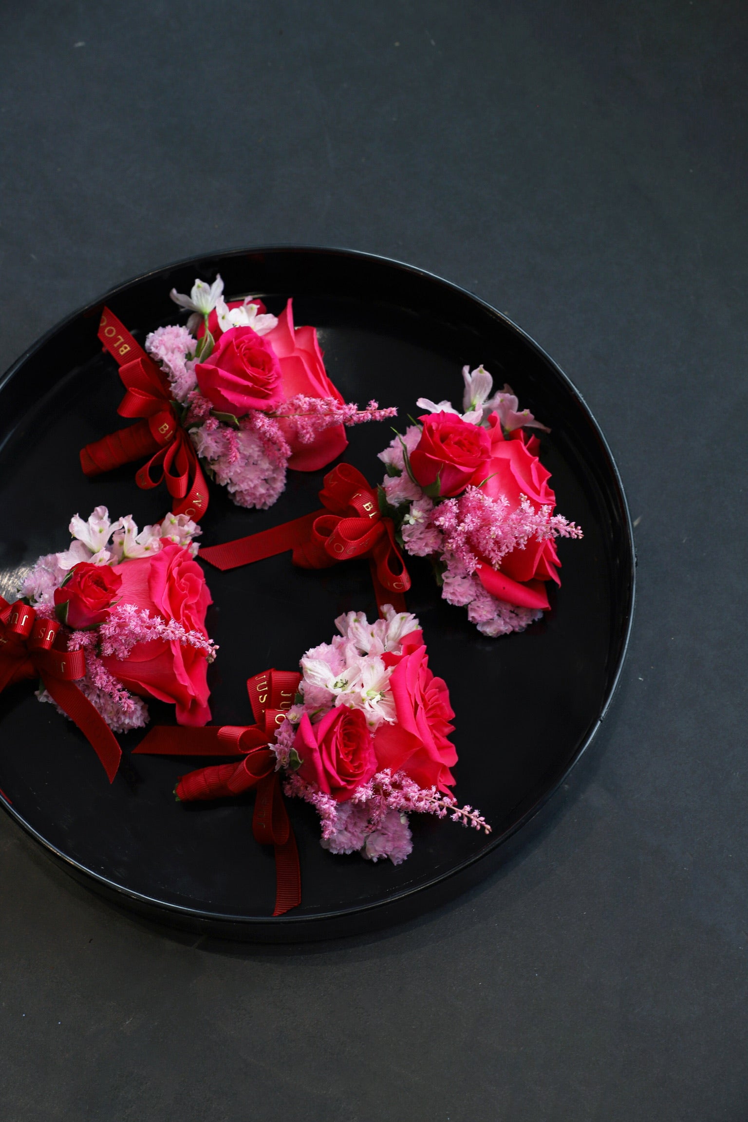 Close-up of four Vivid Promise wedding corsages on a black tray by Just Bloom Hong Kong, showcasing fuchsia roses, pink astilbes, sweet peas and myosotis with Just Bloom deep red ribbon