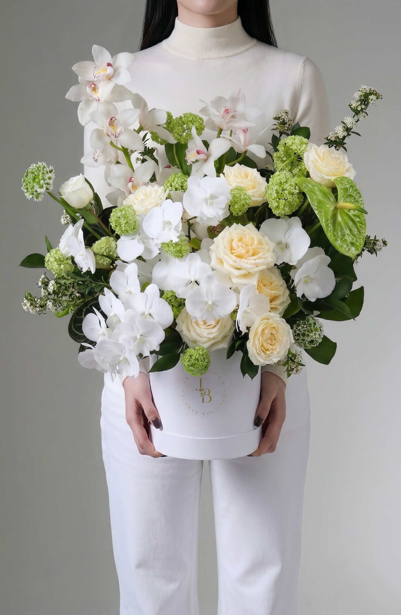 A model in a white outfit holding the White Affair luxury flower box by Just Bloom Hong Kong, showcasing white orchids, cream roses, green viburnum and anthuriums in a white hat box