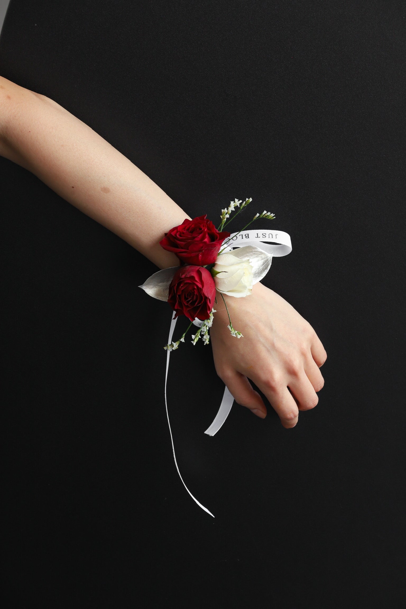 A model wearing Wrist Corsage No.4 featuring deep red roses, a white rose, seasonal fillers and silver foliage tied with a Just Bloom white ribbon — Just Bloom Hong Kong