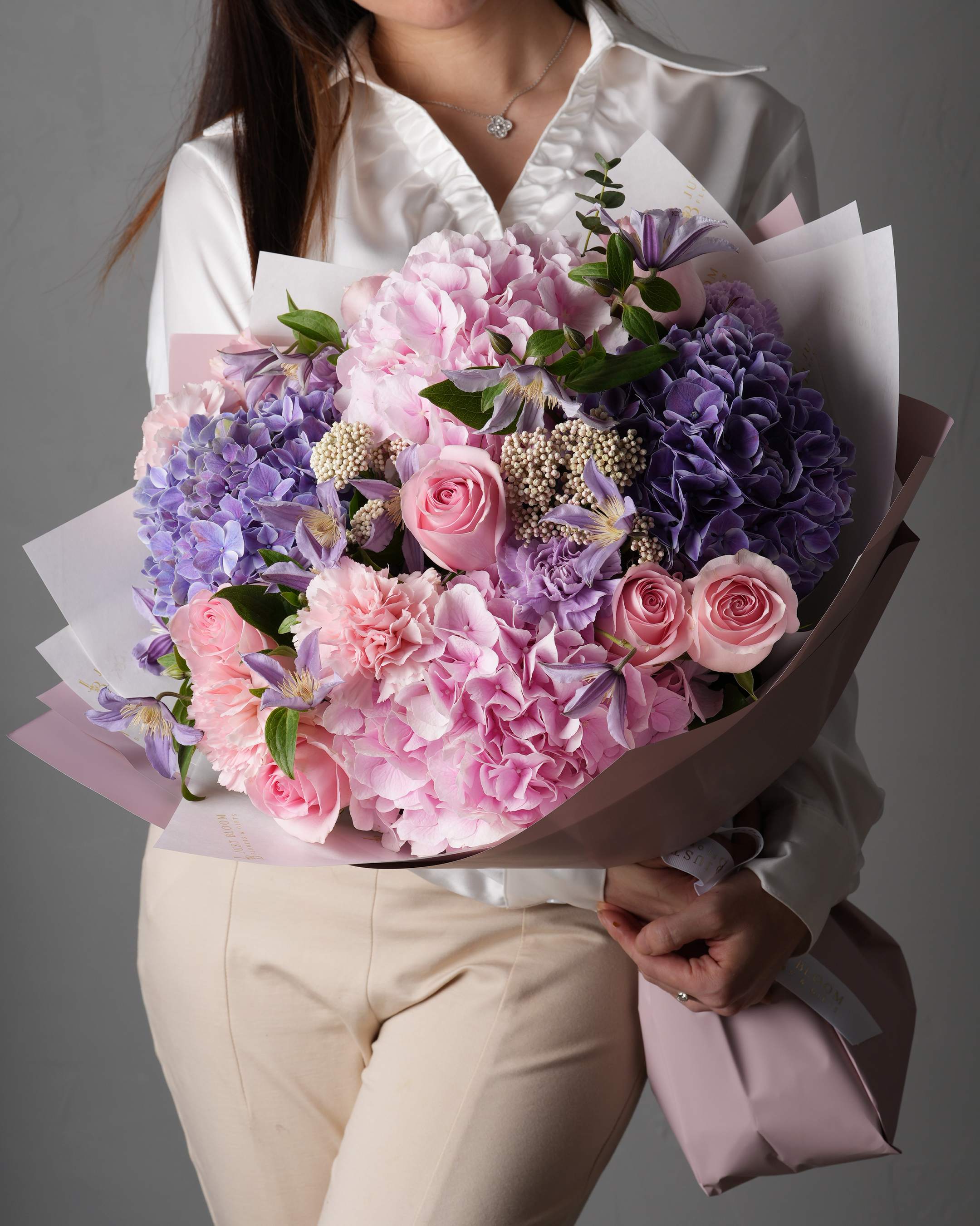 Woman in a white blouse holding a large bouquet of pink and purple hydrangeas, roses, carnations, clematis, and rice flowers.