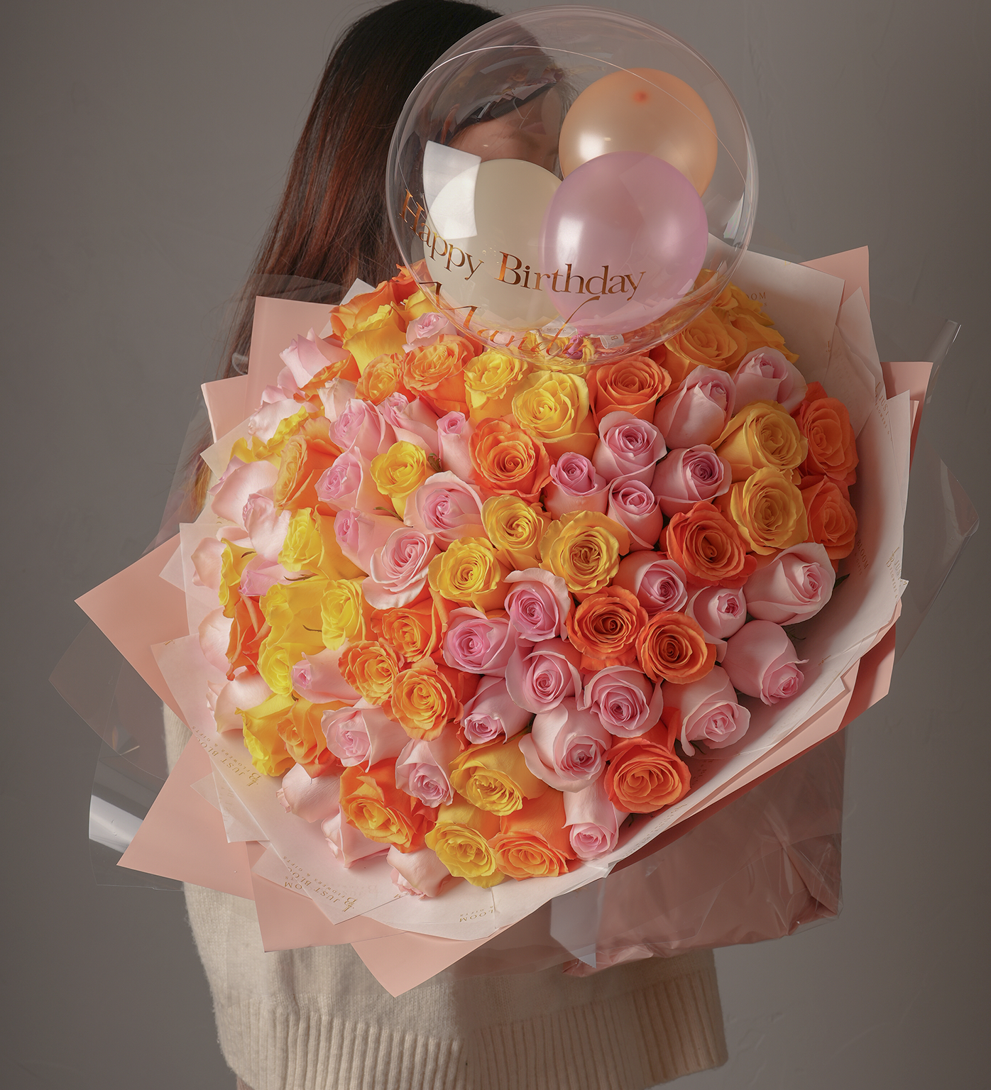 Woman holding large 99-stem bouquet with pink, yellow, and orange Ecuadorian roses, topped with a “Happy Birthday” crystal balloon