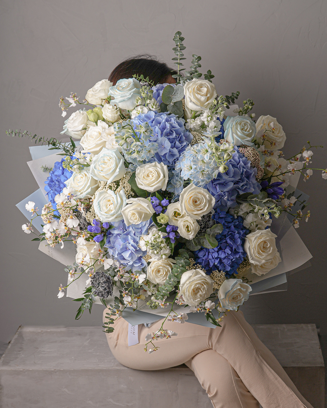 Emma bouquet featuring white roses, blue and lavender hydrangeas, white stock, and fillers in soft blue wrapping